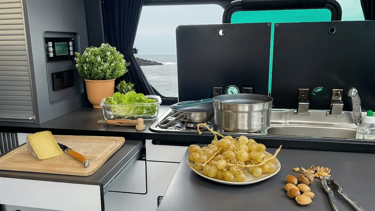 La cuisine du van : deux feux, évier, tiroir plan de travail, avec vue sur la mer par la fenêtre