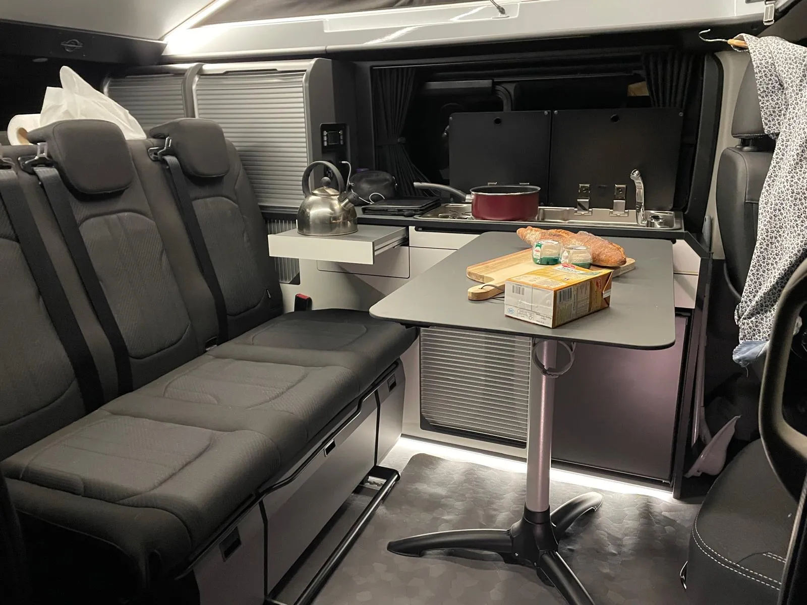 L’intérieur du van : banquette, table sur pied, cuisine avec plaque de cuisson et évier, éclairage LED au sol